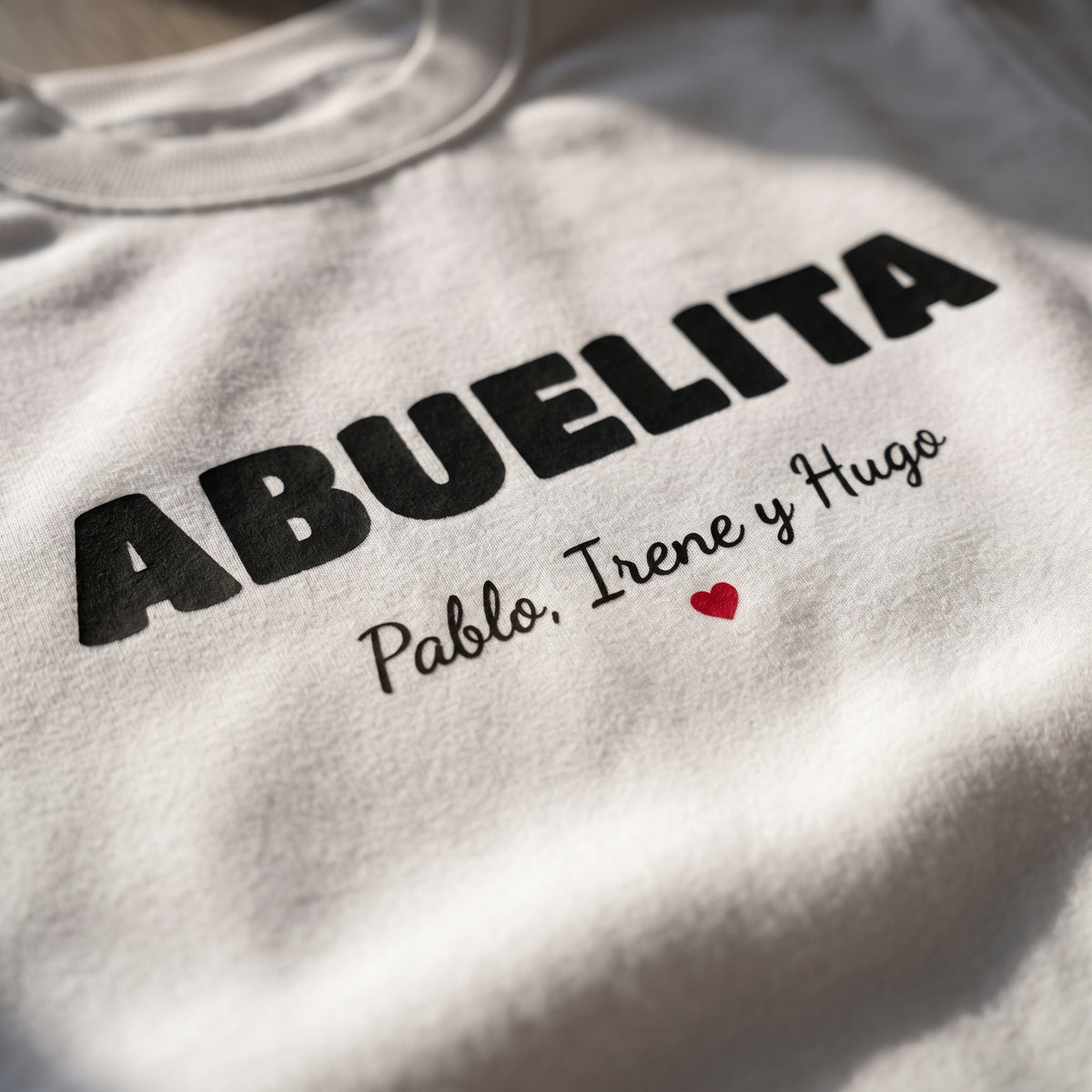 Detalle del estampado DTF de ABUELITA en letra retro con los nombres de los nietos y un corazón rojo sobre una sudadera blanca