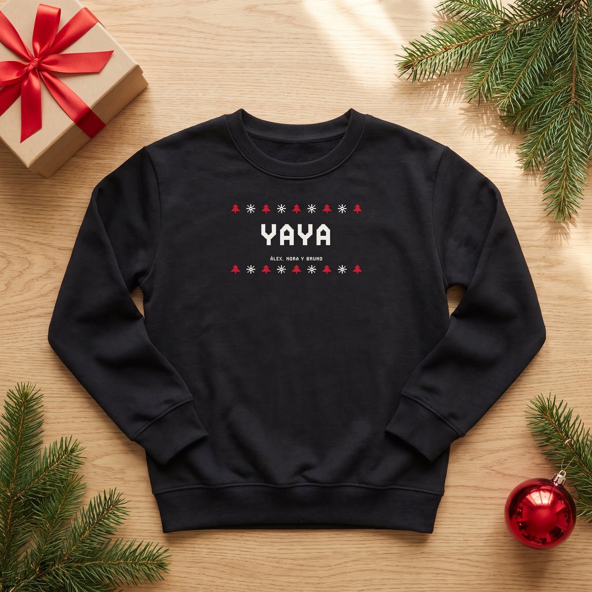 Sudadera negra personalizada con YAYA en estilo jersey navideño de punto, abetos y copos, y los nombres de los nietos, vista de plano junto a un regalo