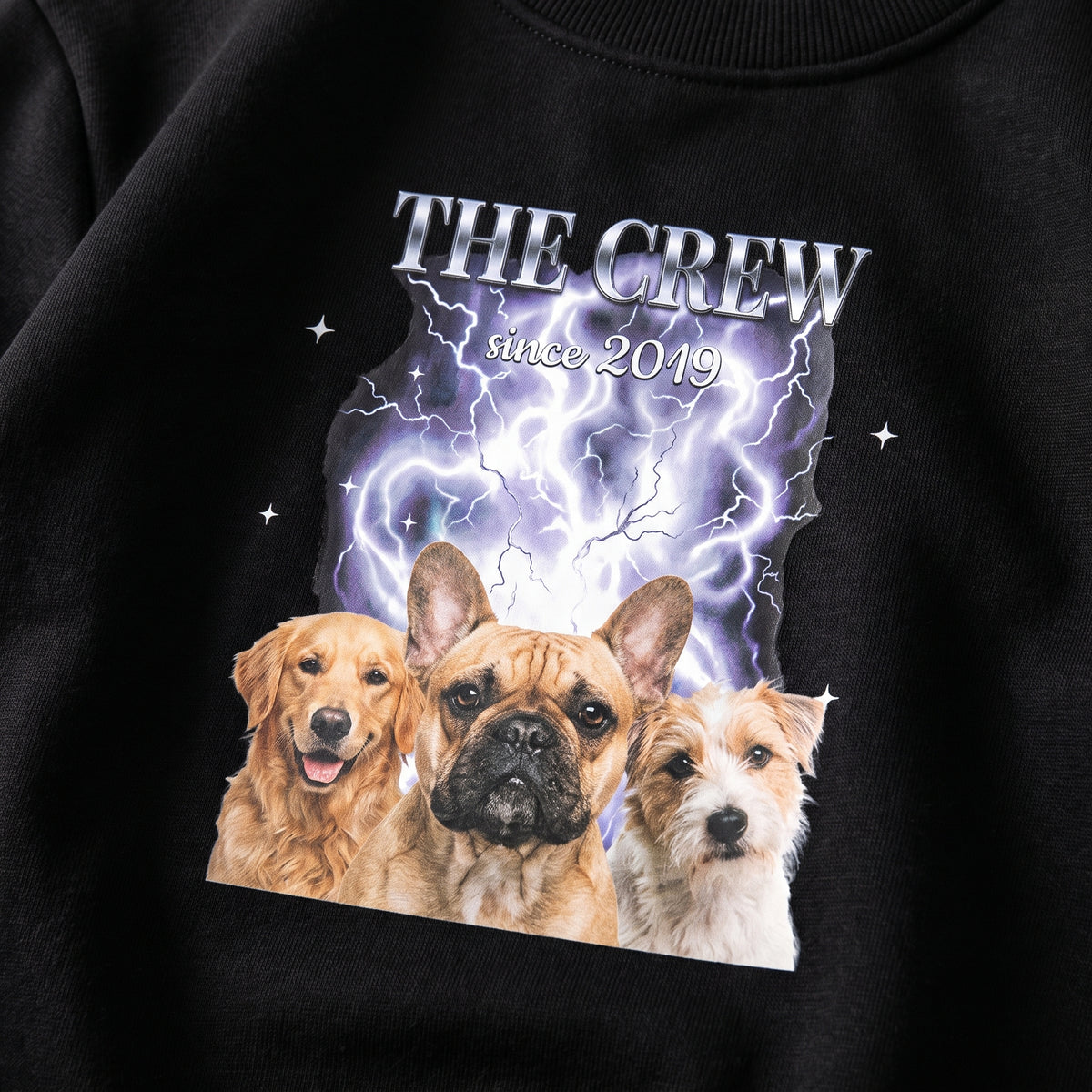 Detalle del estampado DTF estilo bootleg en una sudadera negra: tres perros sobre humo y rayos plateados, el título THE CREW en cromado y «since 2019»