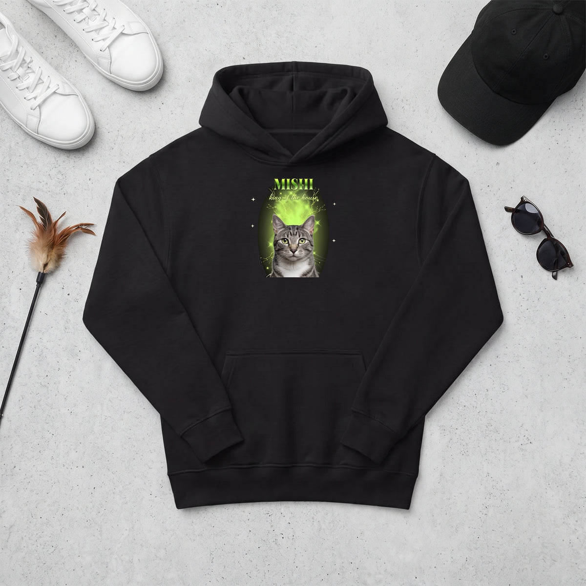 Sudadera con capucha negra estilo bootleg con un gato atigrado sobre humo verde, MISHI y «king of the house», junto a unas zapatillas, una gorra y un juguete de gato