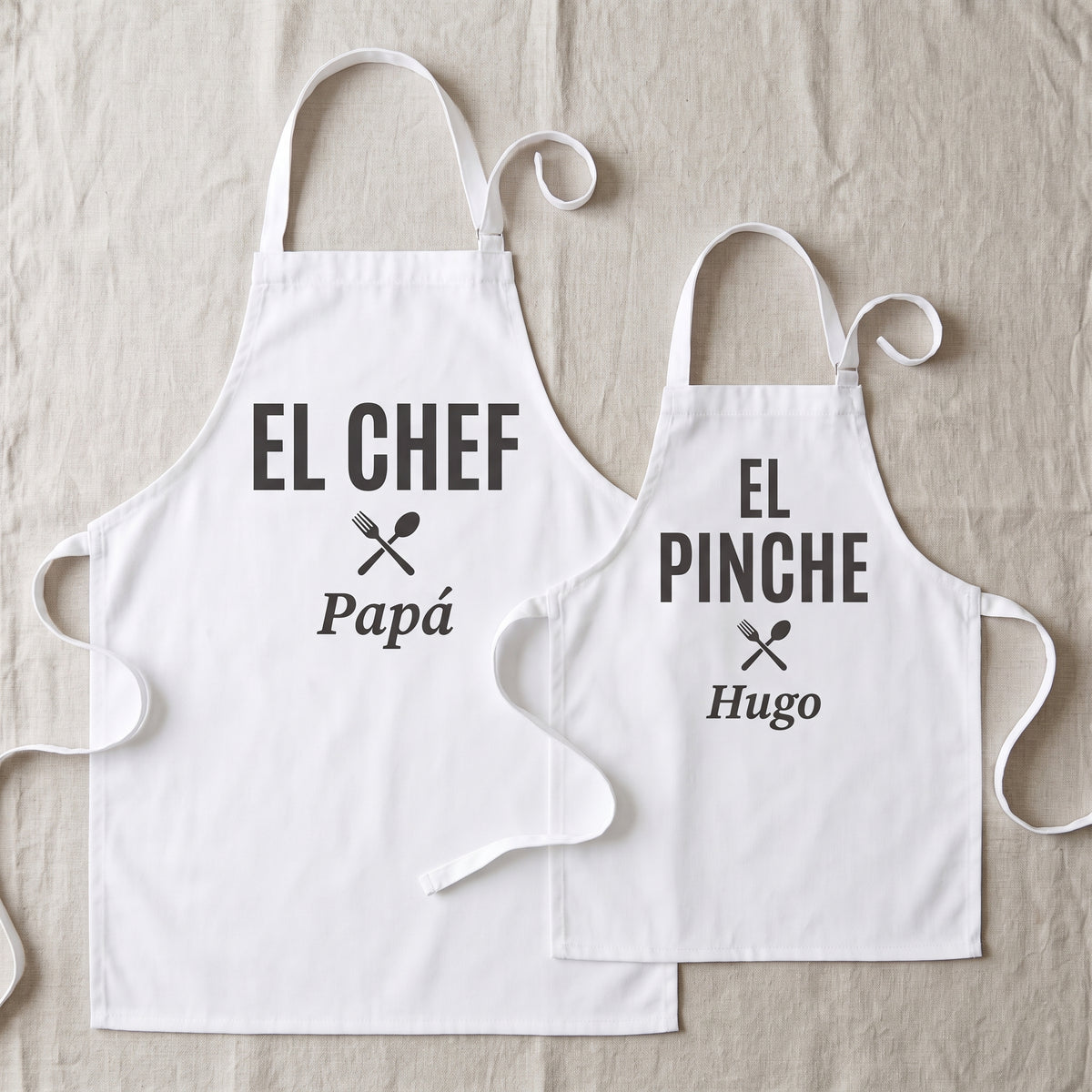 Dos delantales blancos a juego extendidos, el de adulto con «EL CHEF» y el nombre Papá y el de niño, más pequeño, con «EL PINCHE» y el nombre Hugo