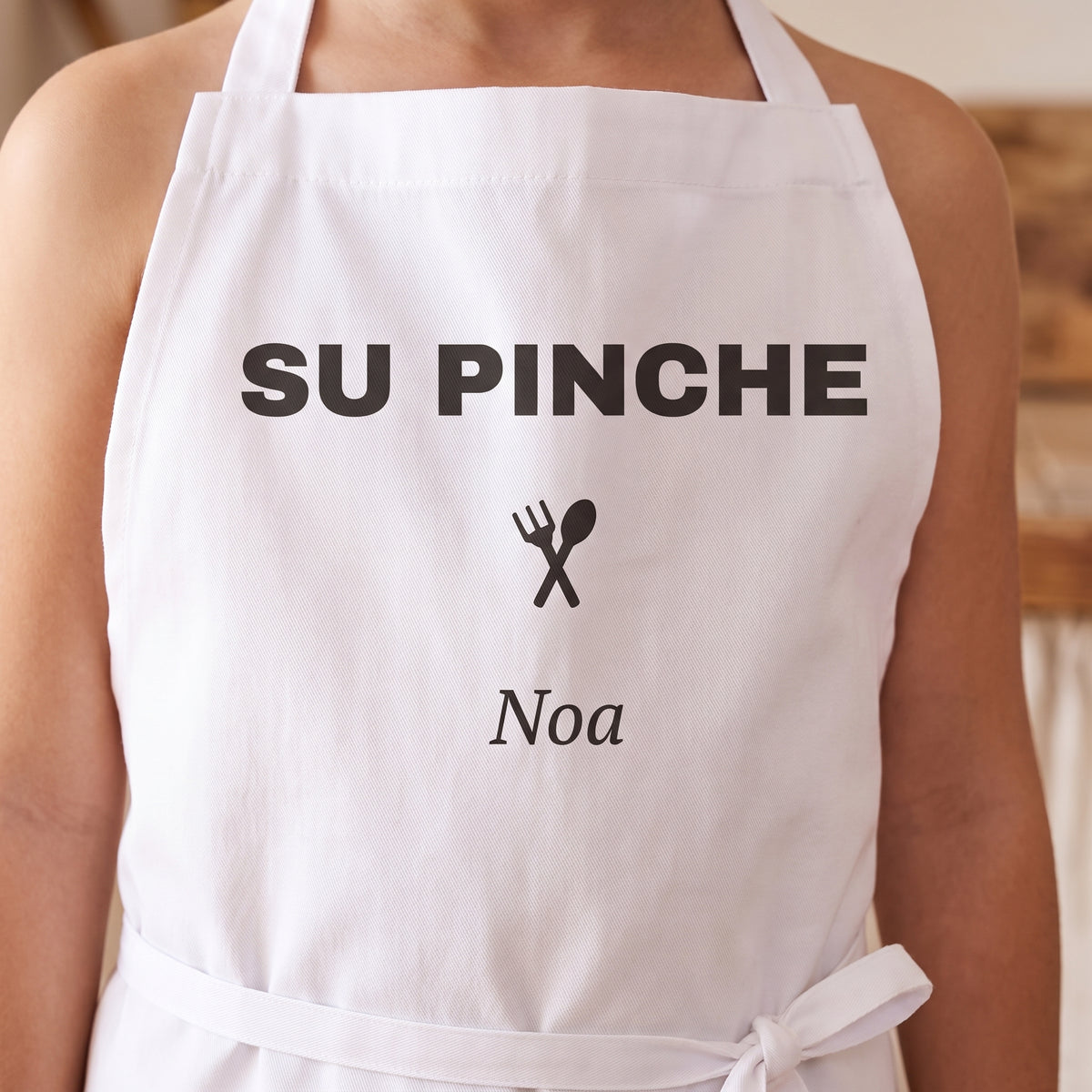 Detalle del peto del delantal pequeño con «SU PINCHE», los cubiertos cruzados y el nombre Noa