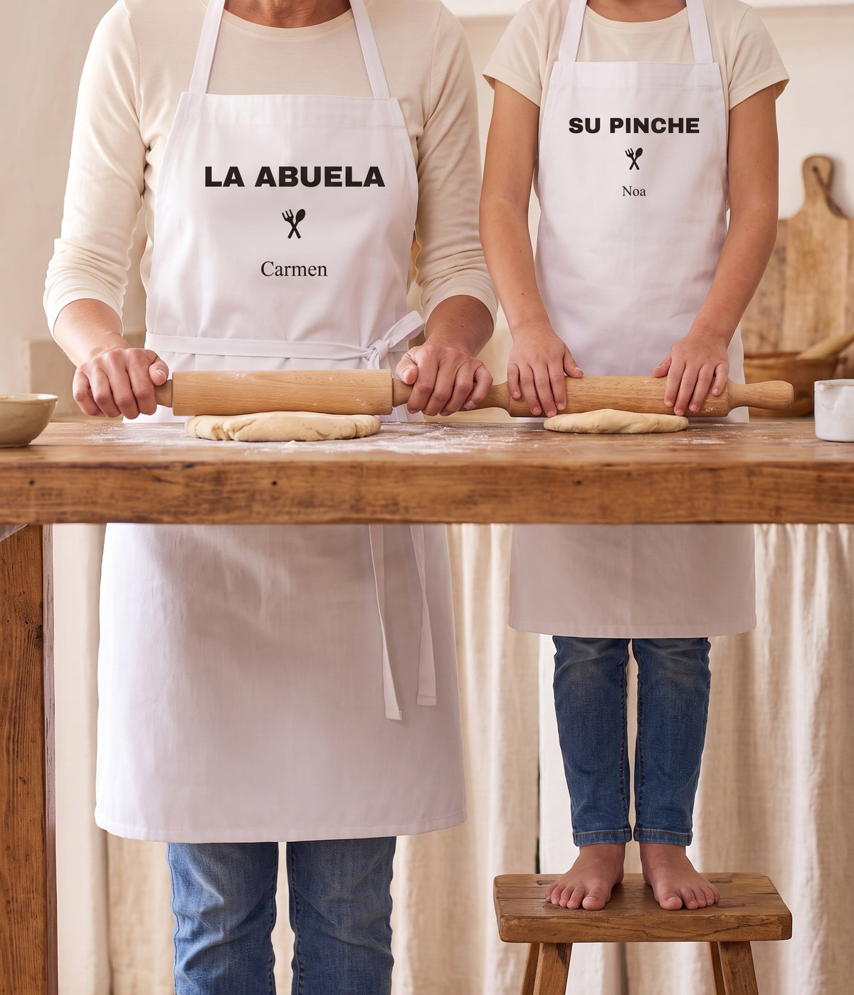 Una abuela y su nieta amasando con los dos delantales personalizados: «LA ABUELA · Carmen» y «SU PINCHE · Noa»