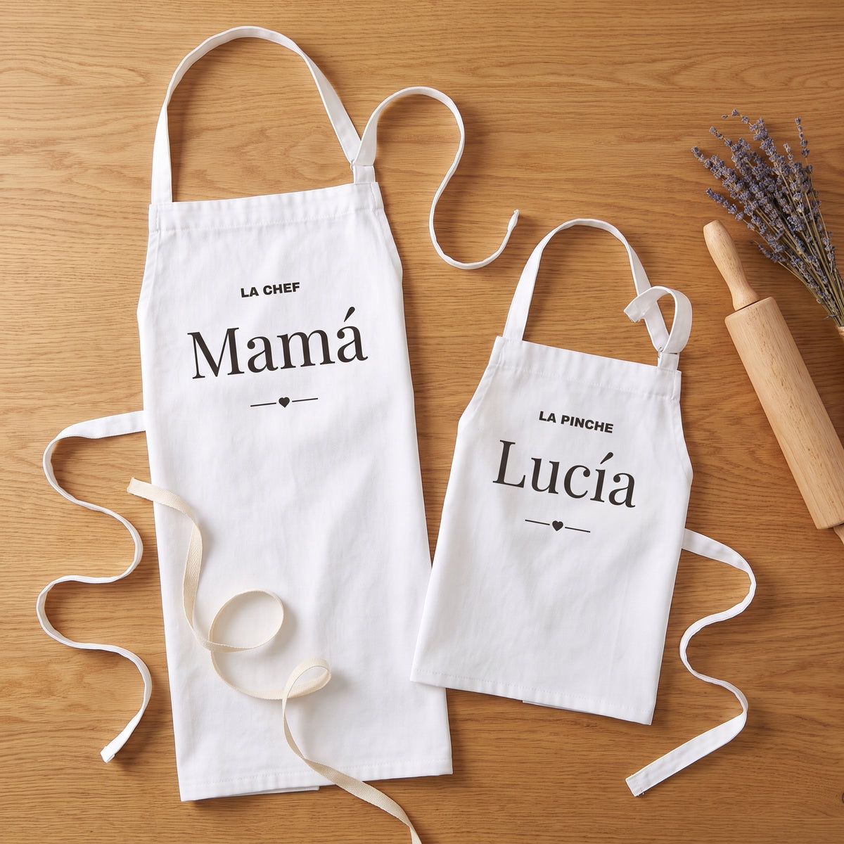 Los dos delantales doblados sobre una mesa de roble con un rodillo, lavanda y una cinta: «LA CHEF · Mamá» y «LA PINCHE · Lucía», listos para regalar