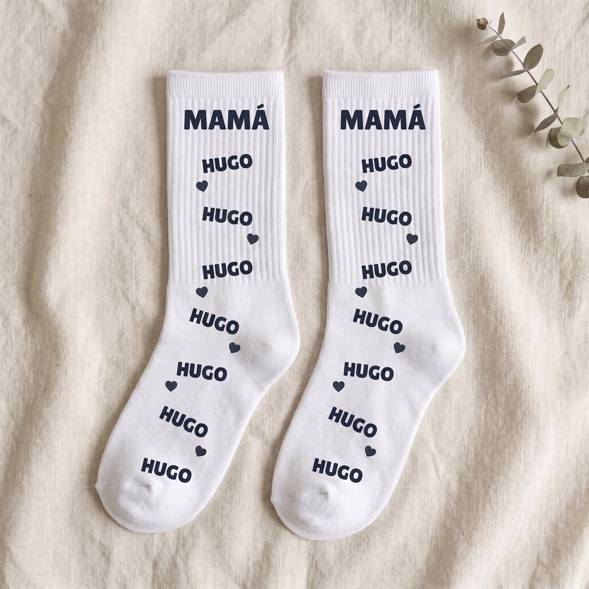 Par de calcetines blancos con MAMÁ en la caña y el nombre HUGO repetido con corazones, vistos de plano