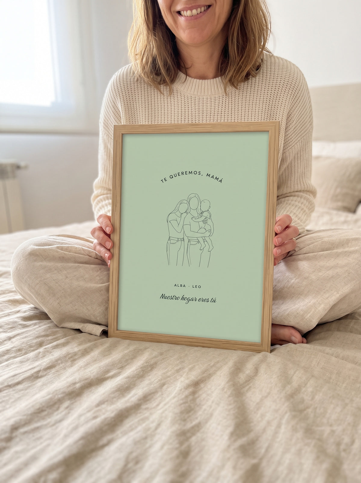 Cuadro mamá de arte lineal personalizado (TE QUEREMOS, MAMÁ · ALBA · LEO · Nuestro hogar eres tú) sostenido entre las manos, con marco de madera
