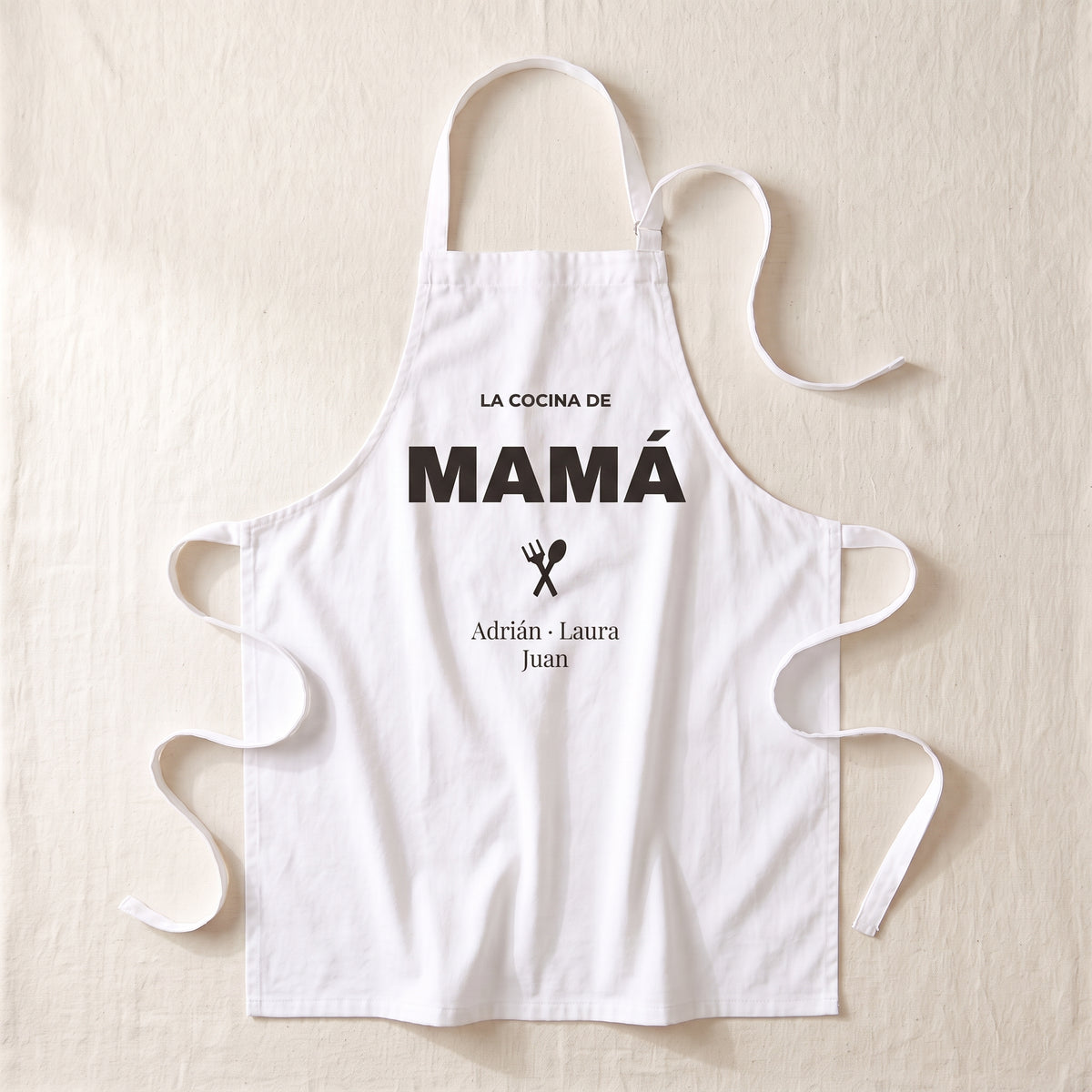 Delantal personalizado blanco extendido, con «LA COCINA DE MAMÁ», un tenedor y una cuchara cruzados y los nombres Adrián, Laura y Juan