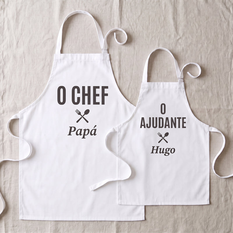 Dos delantales blancos a juego extendidos, el de adulto con «EL CHEF» y el nombre Papá y el de niño, mais pequeño, con «EL PINCHE» y el nombre Hugo