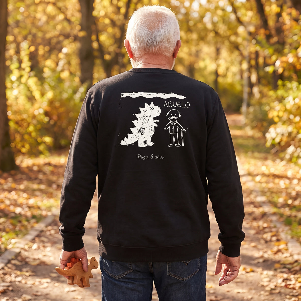 Abuelo de espaldas paseando por un parque en otoño con una sudadera negra personalizada con el dibujo de su nieto: un dinosaurio, el abuelo y «ABUELO», firmado «Hugo, 5 años»