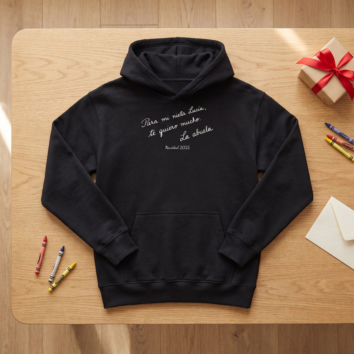 Sudadera con capucha negra personalizada con una nota escrita a mano, «Para mi nieta Lucía, te quiero mucho. La abuela», junto a unas ceras y un regalo