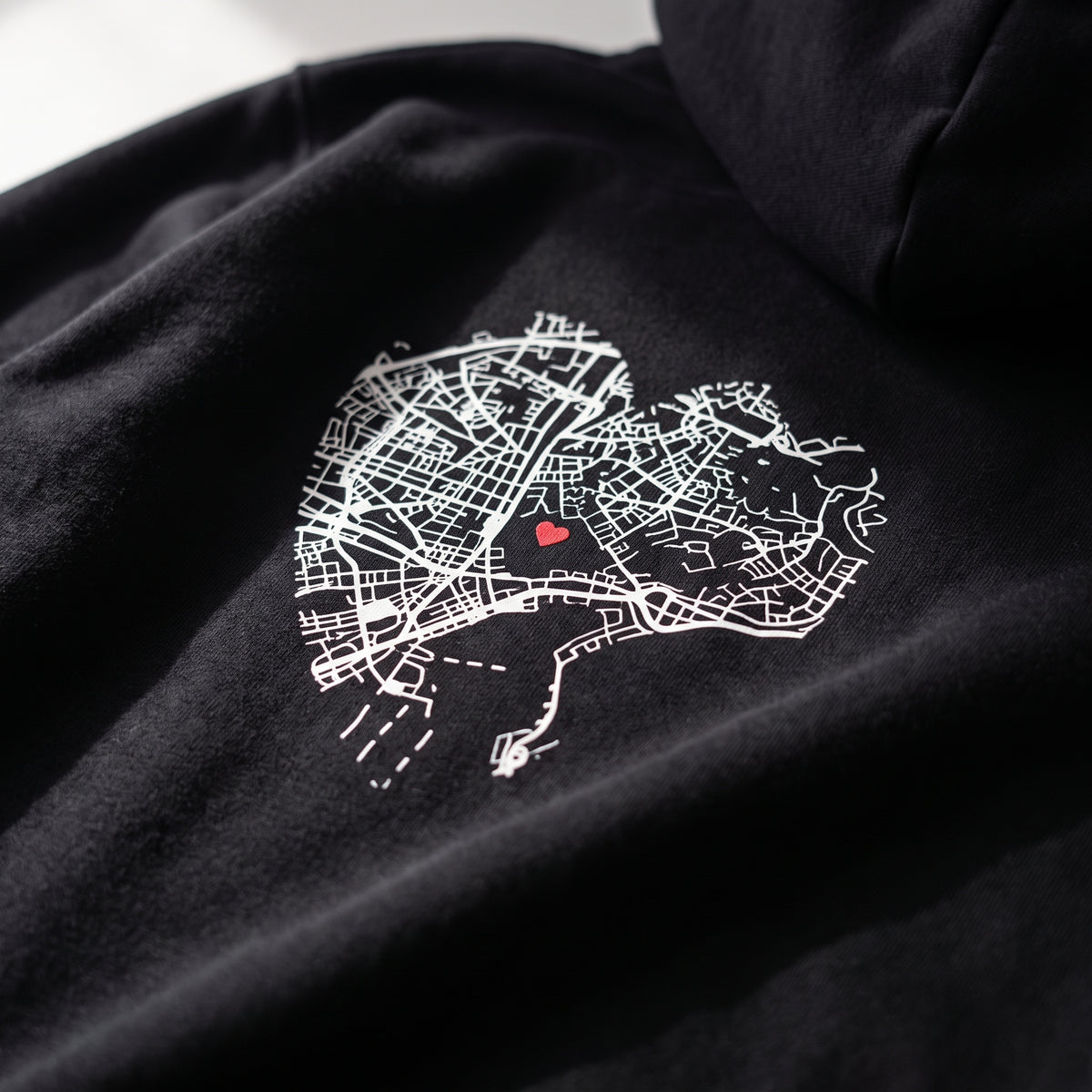 Detalle del estampado DTF de los mapas en corazón de Málaga y Lisboa sobre el tejido de una sudadera con capucha negra