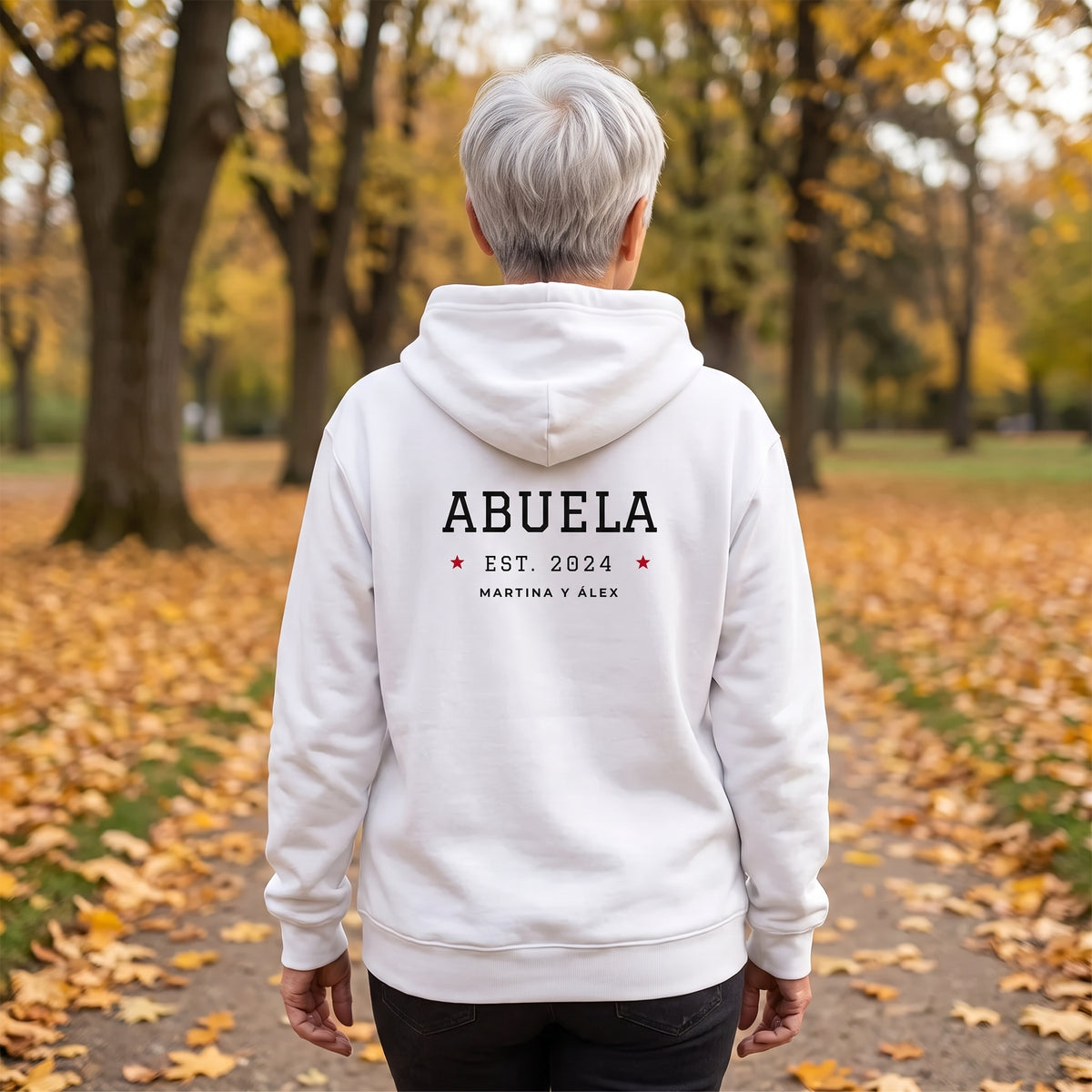 Mujer de espaldas en un parque en otoño con la sudadera con capucha blanca «ABUELA EST. 2024», Martina y Álex, estilo universitario en la espalda