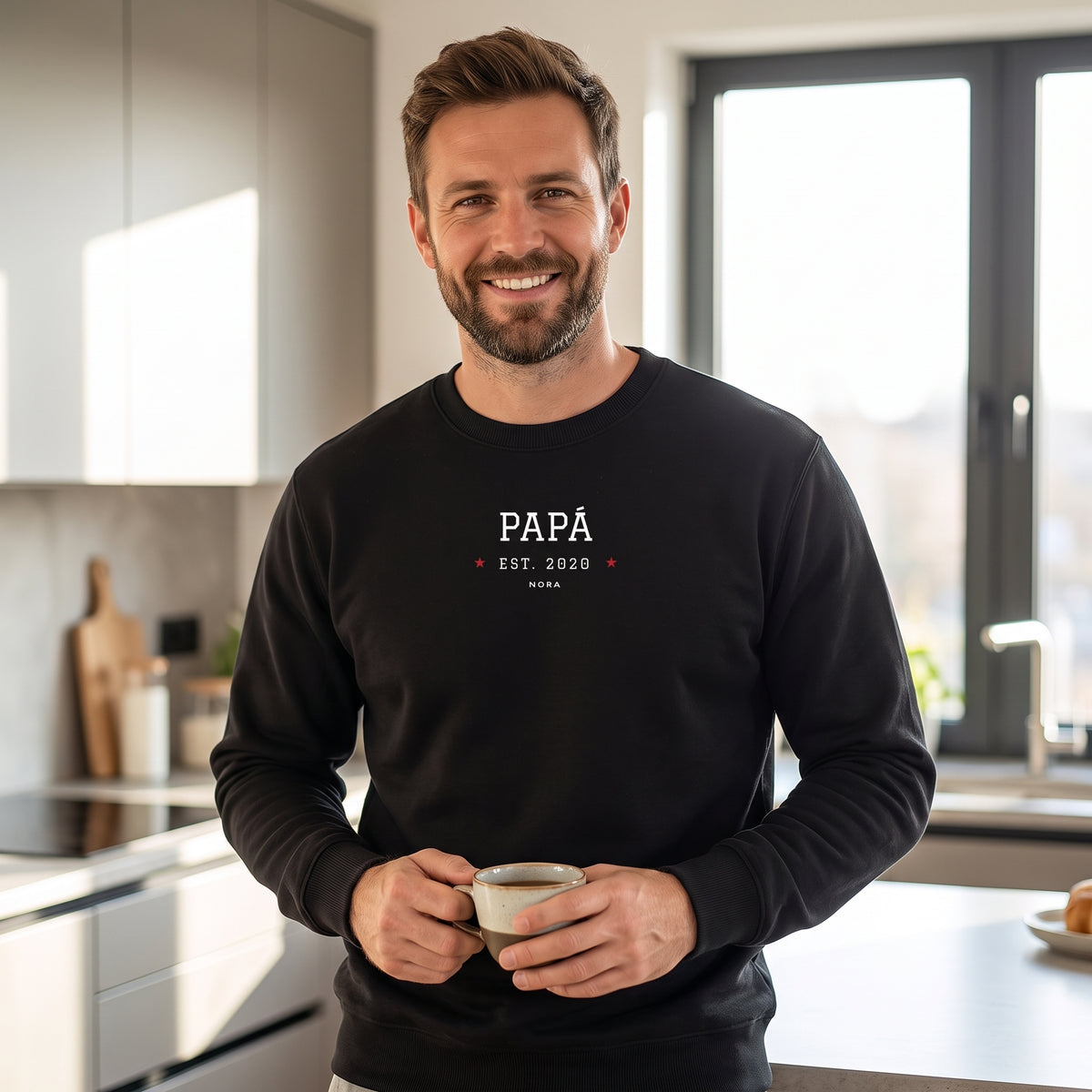 Hombre en la cocina con una taza de café y la sudadera negra «PAPÁ EST. 2020», Nora, estilo universitario entre dos estrellas rojas
