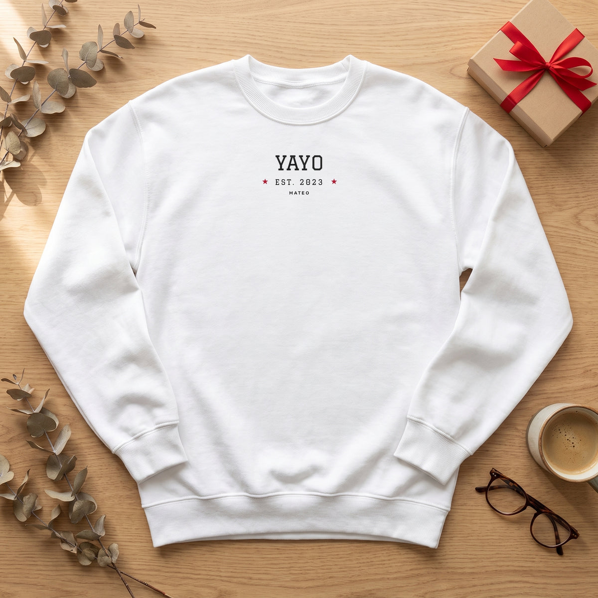 Sudadera blanca «YAYO EST. 2023», Mateo, sobre una mesa de madera con una caja de regalo, café y unas gafas
