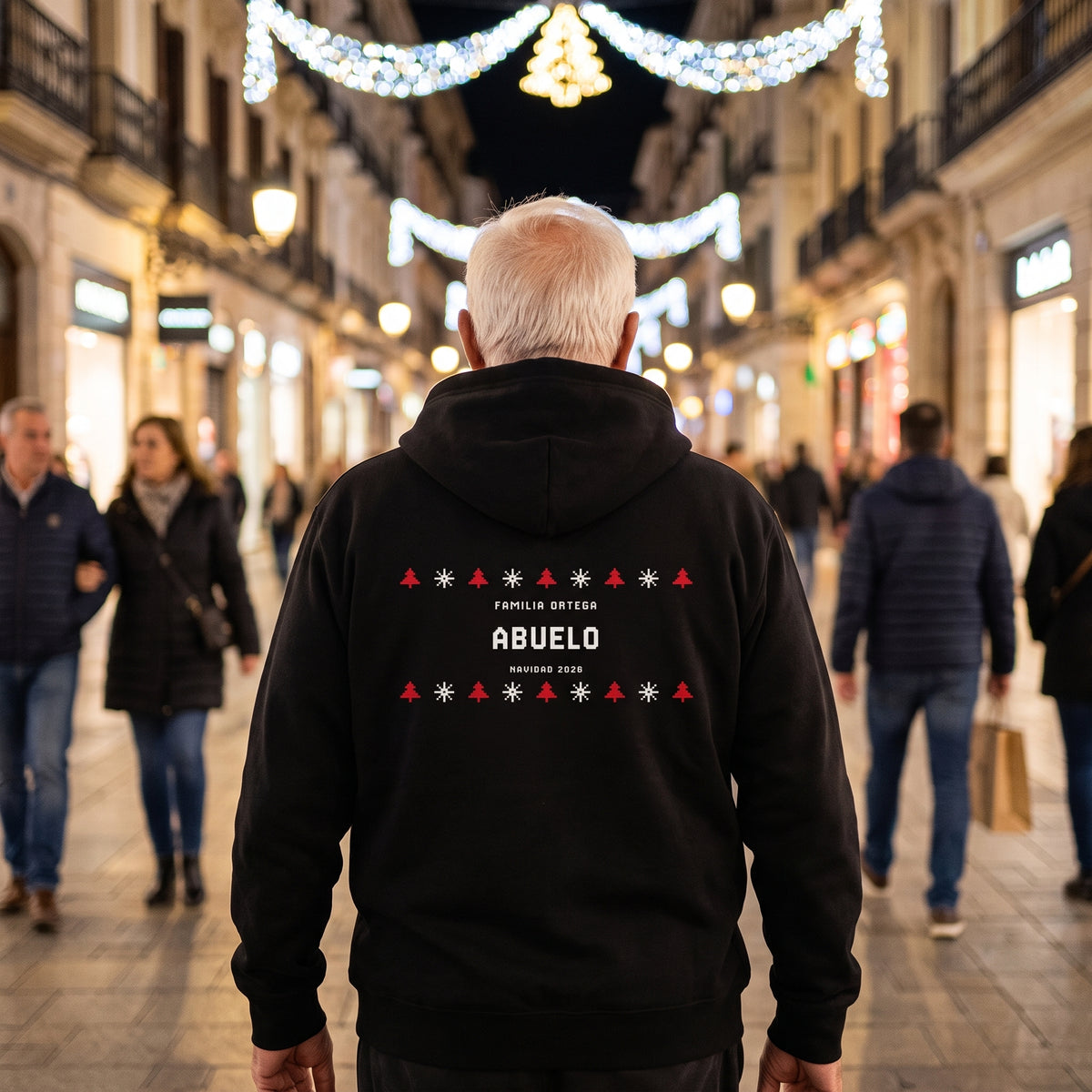Hombre mayor de espaldas en una calle con luces de Navidad con la sudadera con capucha negra estilo jersey de punto «FAMILIA ORTEGA · ABUELO · NAVIDAD 2026» en la espalda