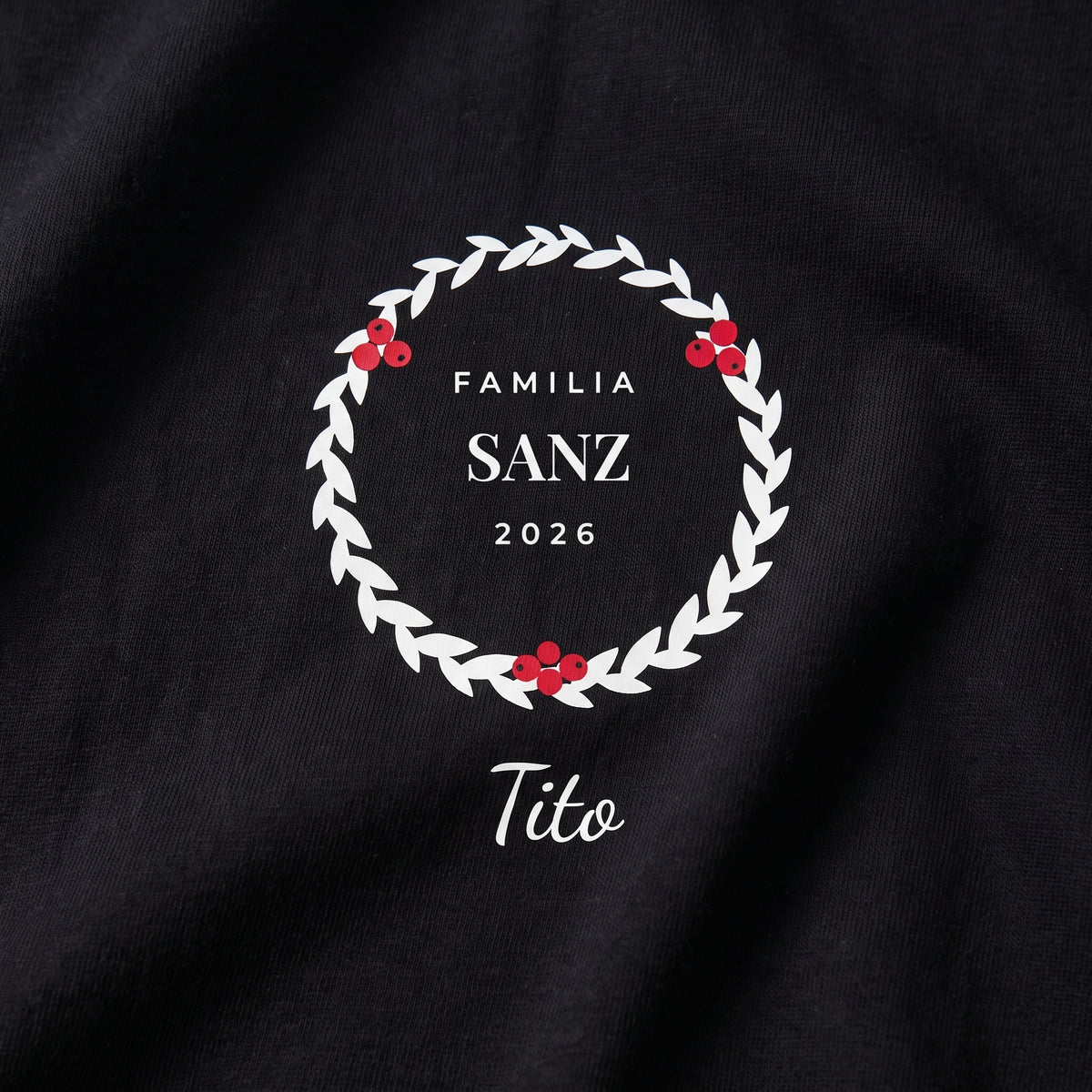 Detalle del estampado DTF sobre una camiseta negra: corona de hojas con bayas rojas, «FAMILIA SANZ 2026» y «Tito» a mano