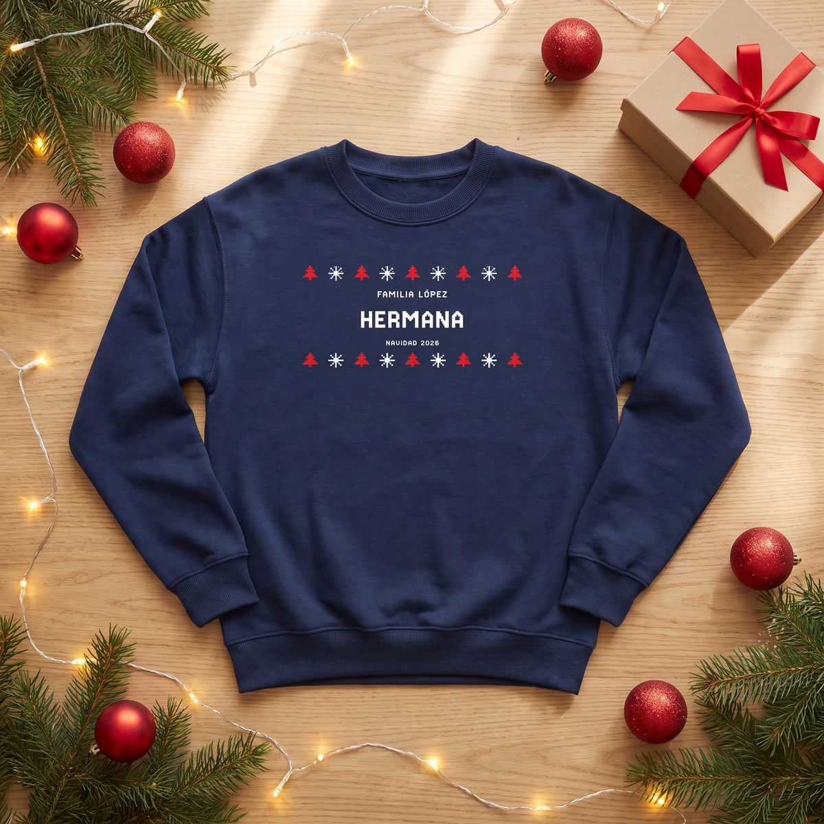 Sudadera azul marino estilo jersey de punto «FAMILIA LÓPEZ · HERMANA · NAVIDAD 2026» sobre una mesa con ramas de pino, bolas rojas y una caja de regalo