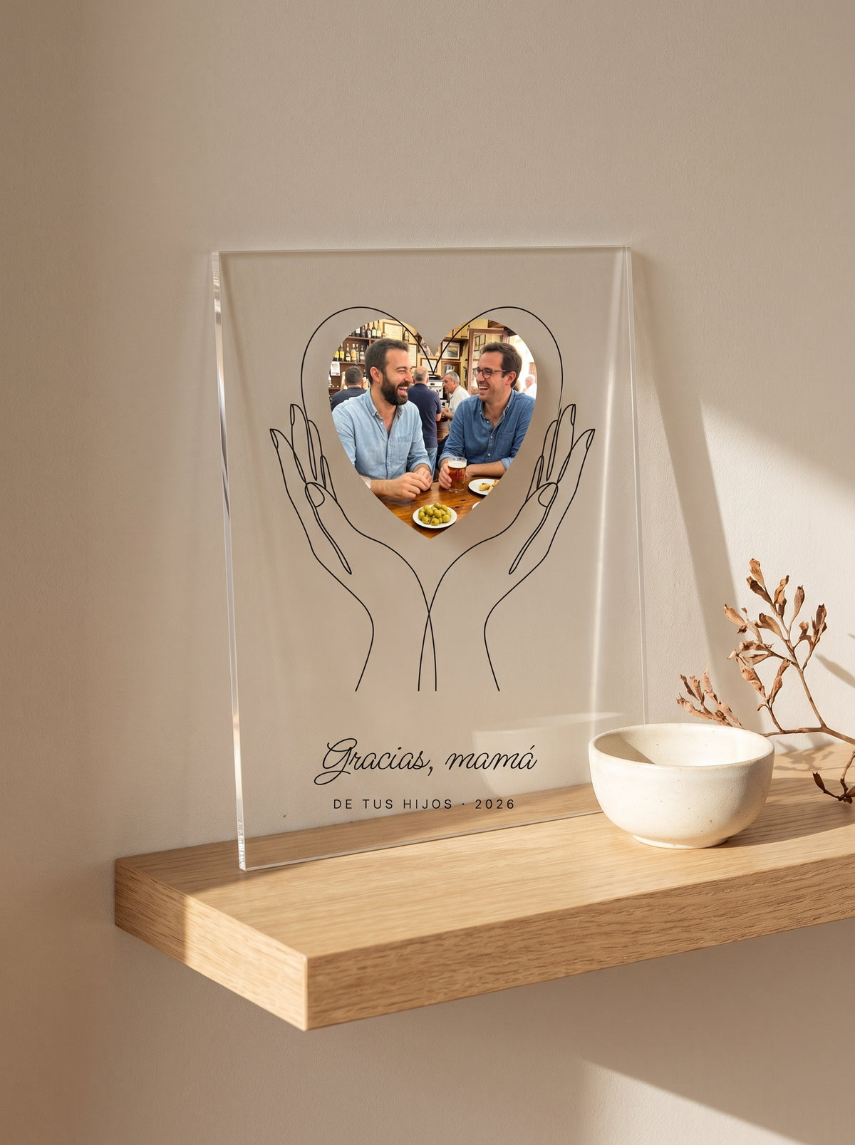 Placa de metacrilato personalizada con unas manos que sostienen la foto apoyada en una balda de roble junto a un cuenco de cerámica y una rama seca