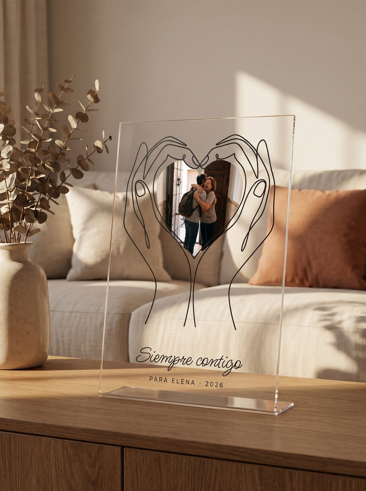 Placa de metacrilato personalizada con unas manos que sostienen la foto sobre el aparador de un salón, con la luz de la tarde entrando por la derecha