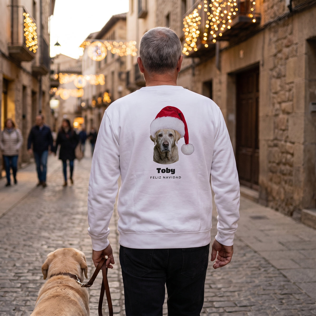 Hombre de espaldas paseando a su labrador por una calle con luces de Navidad, con la sudadera blanca personalizada con la cara del perro con gorro de Papá Noel, Toby y «Feliz Navidad» en la espalda