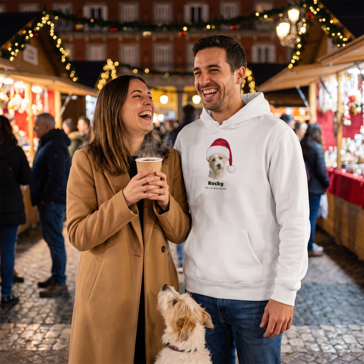 Pareja en un mercado de Navidad con su terrier; él lleva la sudadera con capucha blanca personalizada con la cara del perro con gorro de Papá Noel, Rocky y «Feliz Navidad»