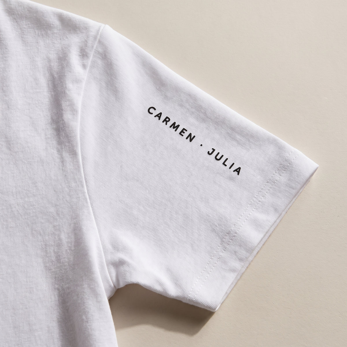 Detalle del estampado DTF a lo largo de la manga de una camiseta blanca: «CARMEN · JULIA» en versalitas espaciadas