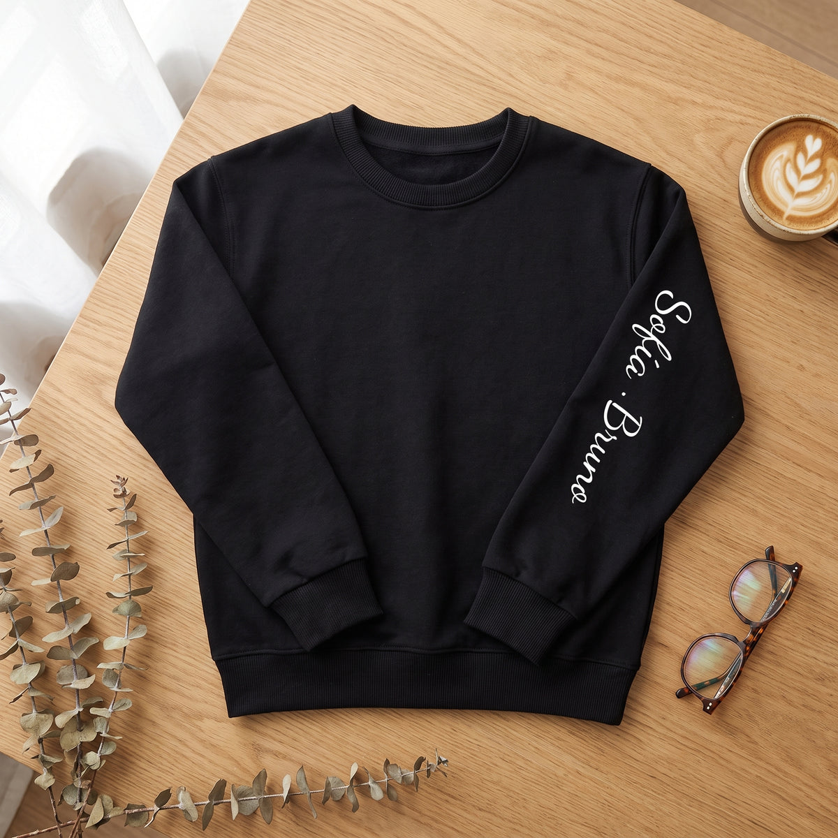 Sudadera negra con «Sofía · Bruno» a mano en la manga, sobre una mesa de madera con un café y unas gafas
