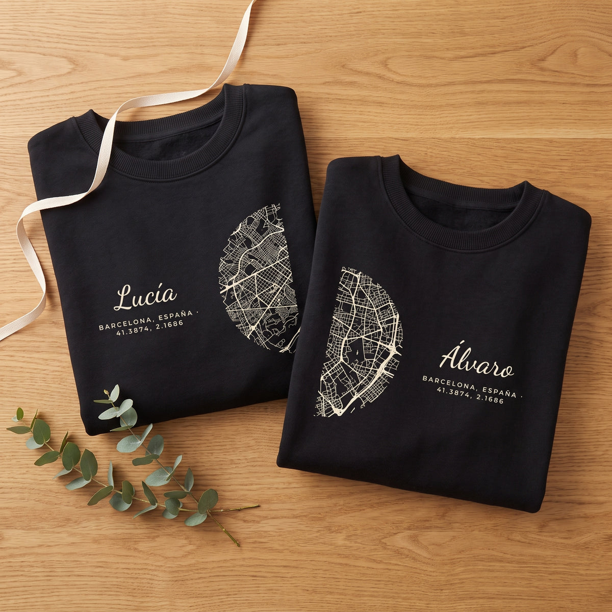 Dos sudaderas negras dobladas sobre una mesa de roble con una cinta y eucalipto: cada una lleva la mitad del mapa de Barcelona, listas para regalar