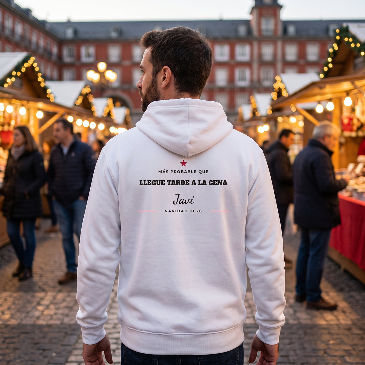 Hombre de espaldas en un mercado navideño con la sudadera con capucha blanca «MÁS PROBABLE QUE LLEGUE TARDE A LA CENA», Javi, estilo diploma, en la espalda