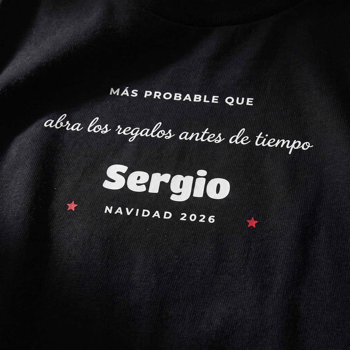 Detalle del estampado DTF sobre una camiseta negra: «MÁS PROBABLE QUE abra los regalos antes de tiempo», Sergio, «NAVIDAD 2026» con dos estrellas rojas