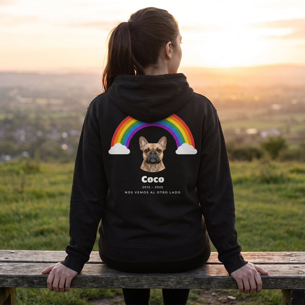 Mujer de espaldas sentada en un banco mirando el atardecer, con una sudadera con capucha negra personalizada con su bulldog francés bajo un arcoíris, Coco, 2012 – 2025 y «Nos vemos al otro lado» en la espalda