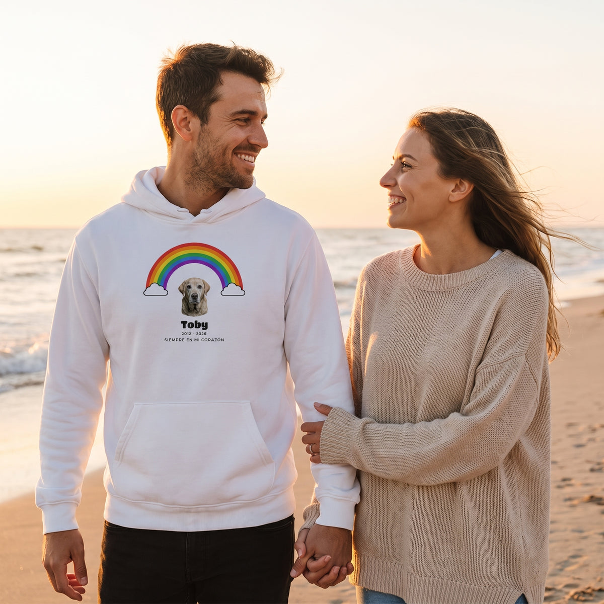 Pareja paseando por la playa al atardecer; él lleva una sudadera con capucha blanca personalizada con su labrador bajo el arcoíris, Toby, 2012 – 2026 y «Siempre en mi corazón»