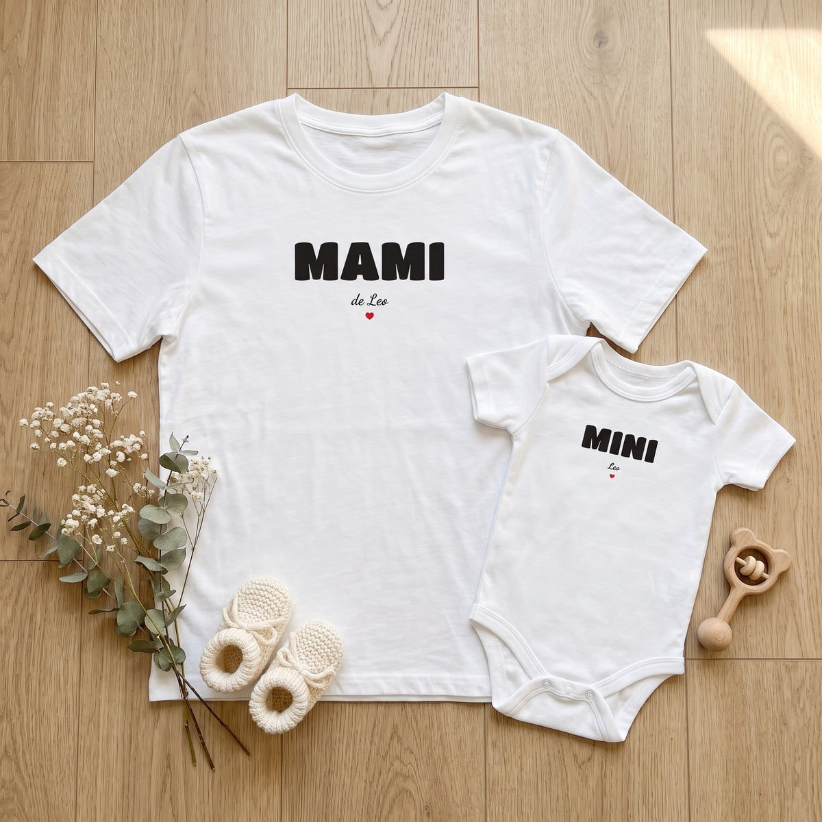 Camiseta blanca «MAMI · de Leo» y body a juego «MINI · Leo» sobre el suelo de madera, con unos patucos de punto y un sonajero