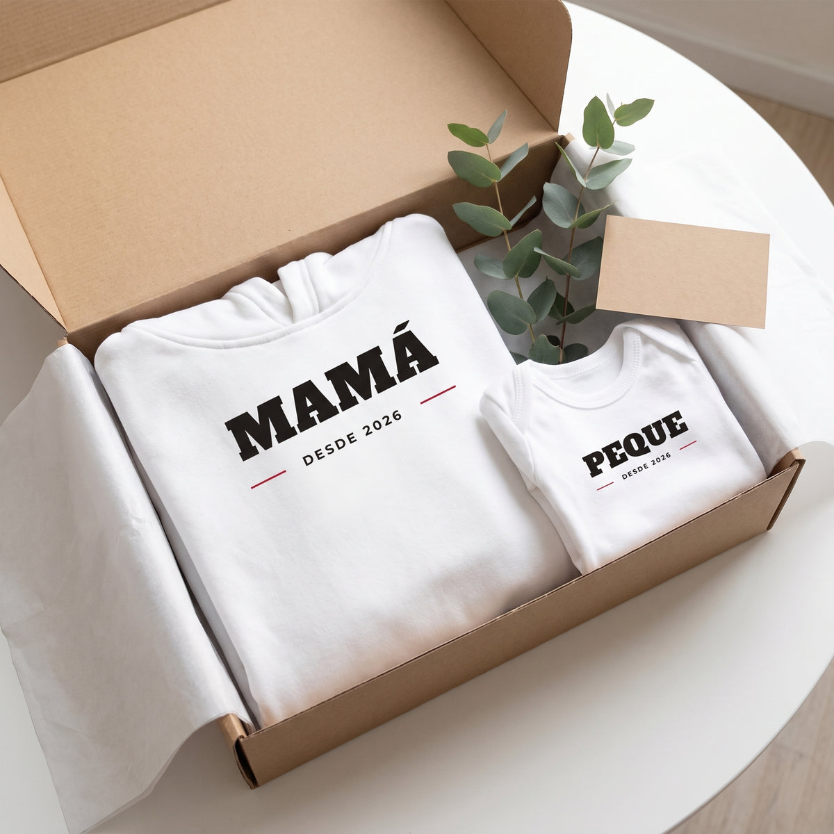 Caja de regalo con la sudadera «MAMÁ · DESDE 2026» y el body «PEQUE · DESDE 2026» doblados