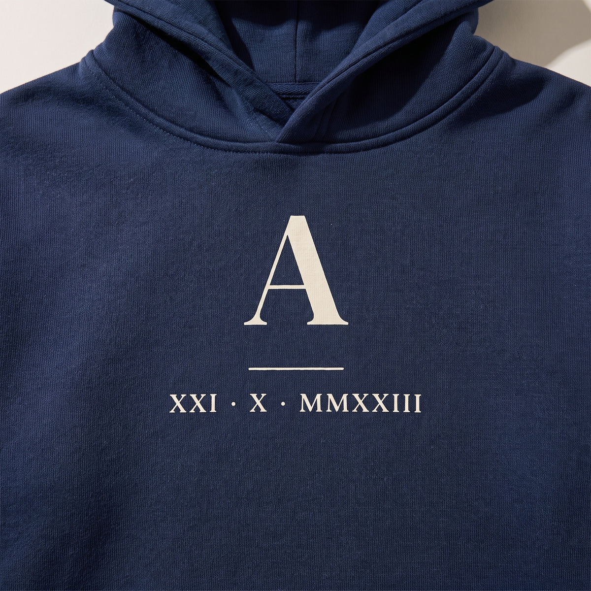 Detalle del estampado en una sudadera con capucha azul marino: la inicial «A» en grande y la fecha en números romanos XXI · X · MMXXIII