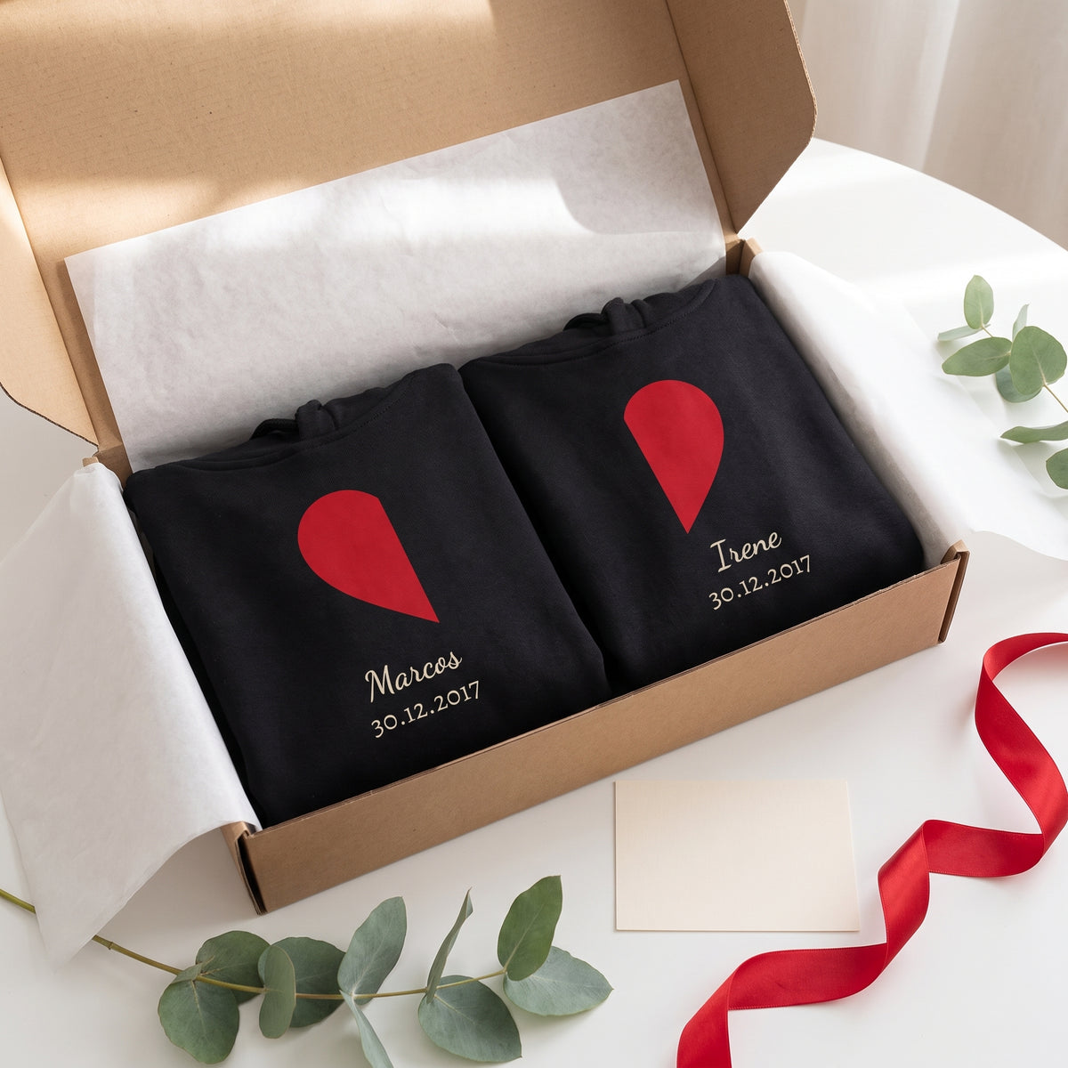 Caja de regalo con dos sudaderas con capucha negras a juego dobladas: medio corazón rojo en cada una, con «Marcos», «Irene» y 30.12.2017