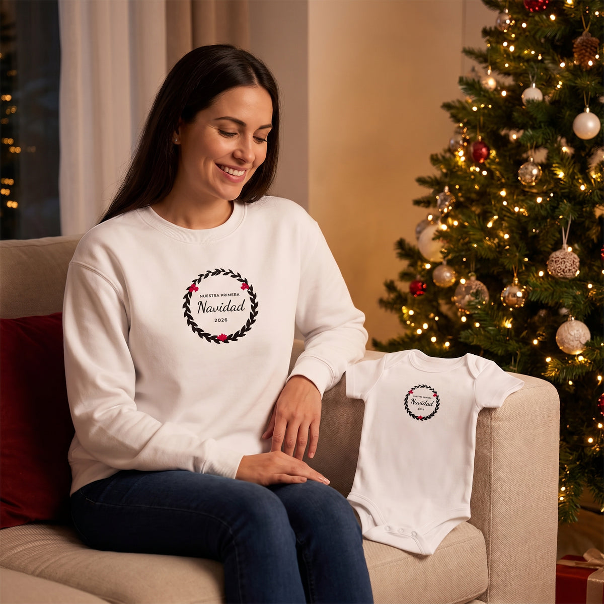 Mamá en el sofá junto al árbol con la sudadera de la corona «NUESTRA PRIMERA Navidad 2026» y el body a juego a su lado