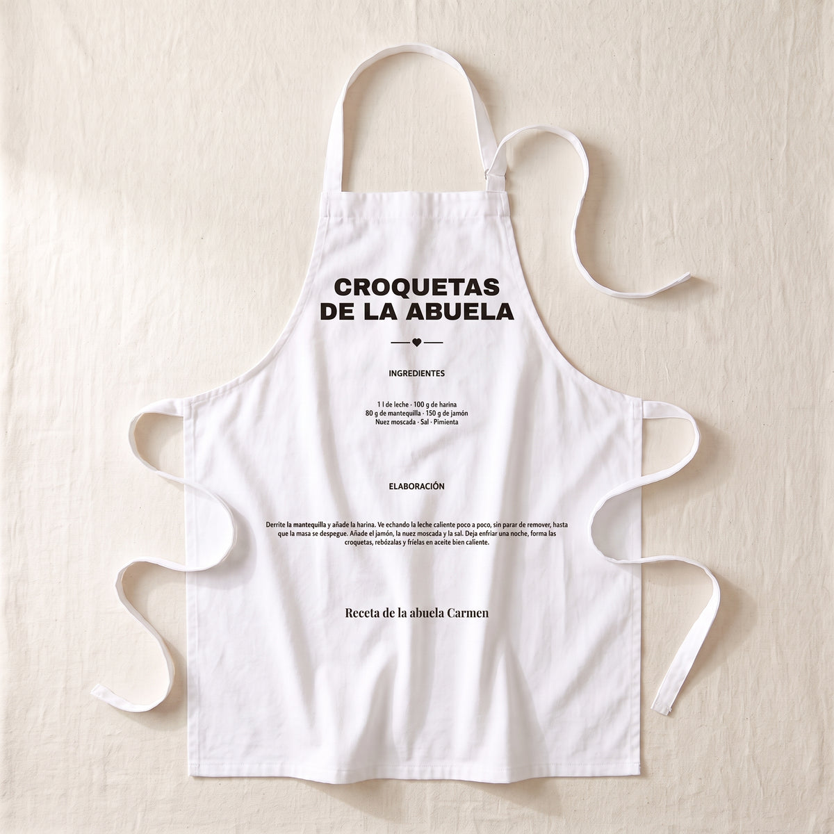 Delantal personalizado blanco extendido, con la receta de las croquetas de la abuela impresa en el peto: el nombre del plato, los ingredientes, la elaboración y la firma «Receta de la abuela Carmen»