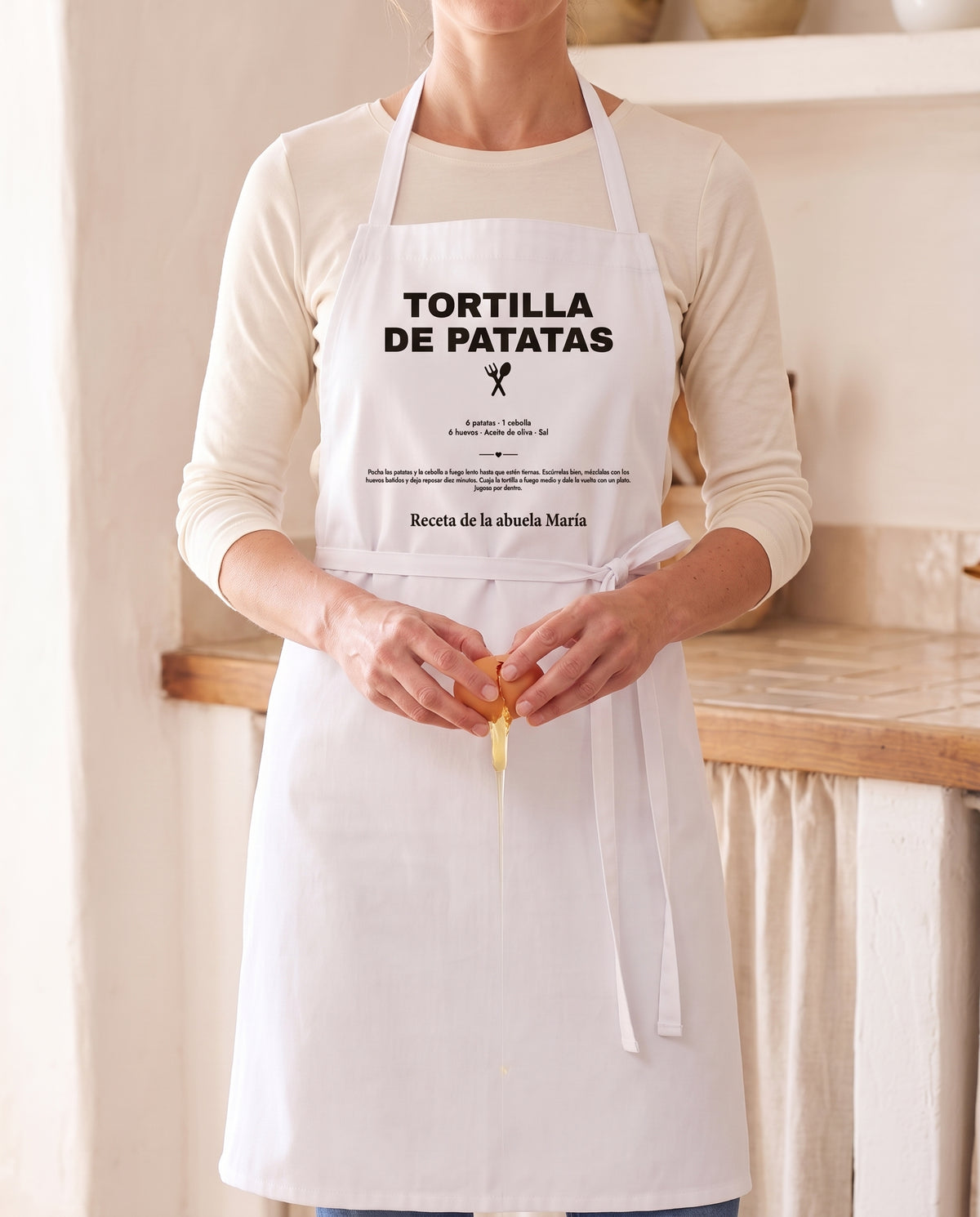 Mujer cascando huevos en la cocina con un delantal personalizado con la receta de la tortilla de patatas de la abuela María