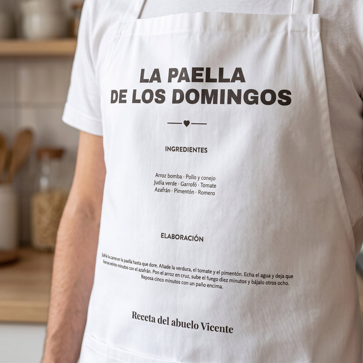 Detalle del peto de un delantal con la receta de «La paella de los domingos», con sus ingredientes y su elaboración en letra pequeña y legible