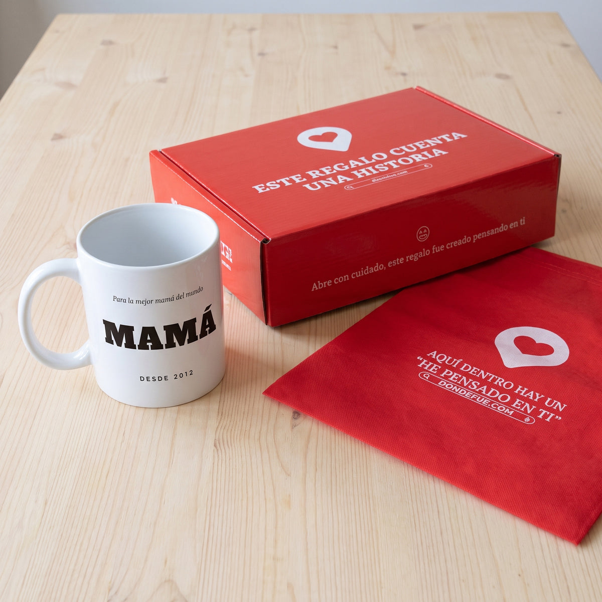 Taza MAMÁ con dedicatoria (Para la mejor mamá del mundo · MAMÁ · DESDE 2012): junto a la caja roja y la bolsa de tela roja en que se envía